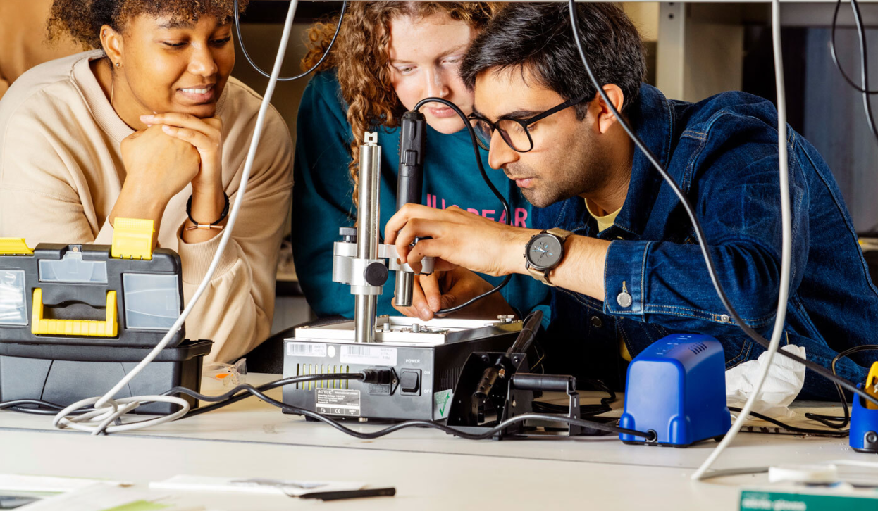 Two MIT undergraduate students smile while observing an instructor working on a circuit board during a hands-on class to build a Bluetooth speaker.