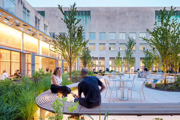 People sit and talk on curved wooden benches in the Hayden Library courtyard, surrounded by trees, greenery, and outdoor tables.