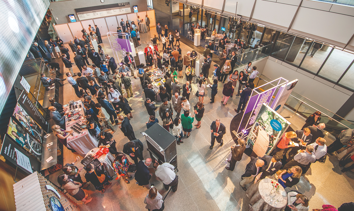 Overhead view of attendees mingling at an MIT event in a large atrium, with display tables and informational booths.