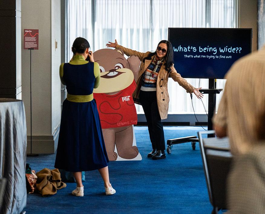 A person photographs a life-size cardboard cutout of the MIT character Wide Tim beside the artist who created him.
